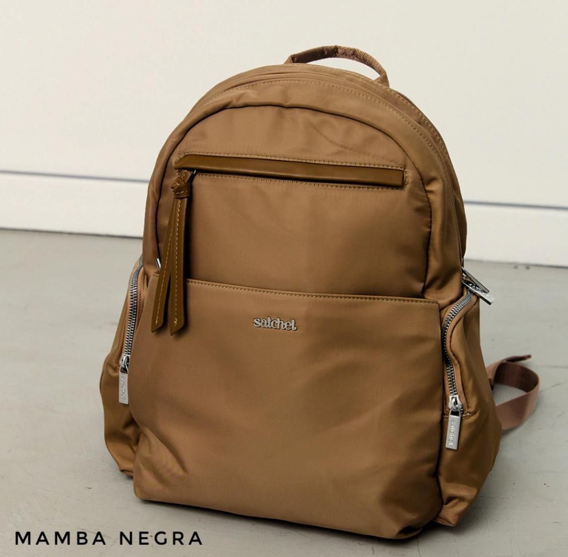 Una mochila de nailon minimalista de color bronce con múltiples bolsillos con cremallera, apoyada contra una pared blanca.