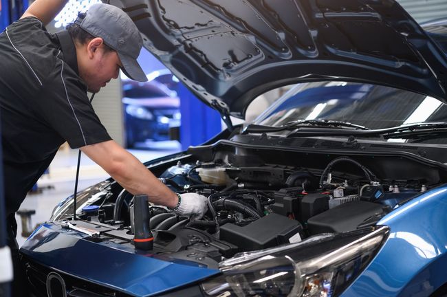 A mechanic wearing a cap and gloves works on the open engine bay of a blue car inside a workshop.