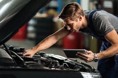 A mechanic in dark coveralls checks a car engine while holding a tablet in a garage.