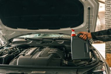 A hand holding a gray jug of motor oil over an open car engine in a garage.