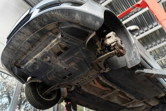 Underside view of a car raised on a lift in a repair shop, showing exposed wheel assembly and protective plastic panels.