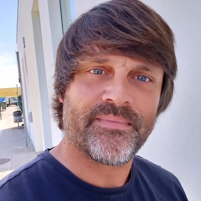 Homem de olhos azuis e barba, vestindo uma camisa azul escura, em pé do lado de fora com uma expressão neutra.
