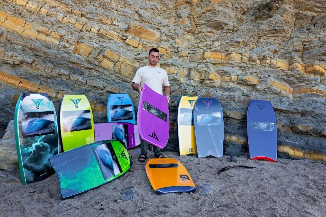 Homem segurando uma prancha de bodyboard roxa, cercado por pranchas de bodyboard de várias cores em uma praia de areia contra um penhasco rochoso.