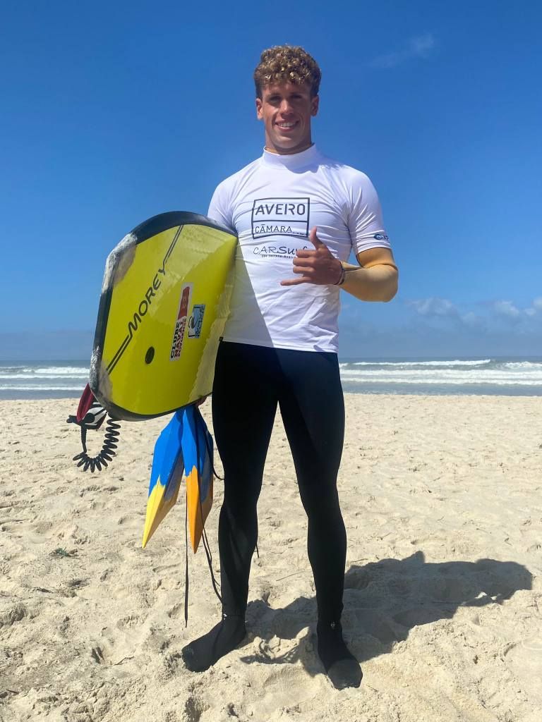 Homem de roupa de mergulho na praia segurando uma prancha de bodyboard e fazendo um sinal de positivo com o polegar.