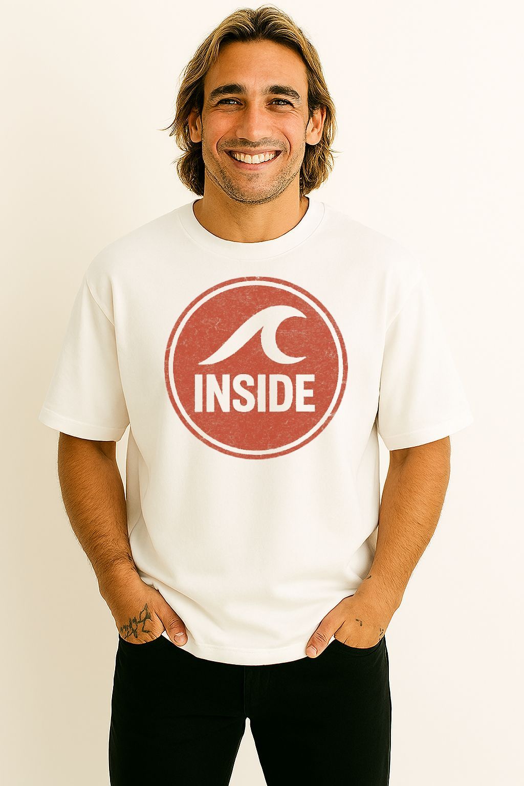 Homem de cabelo comprido, vestindo uma camisa creme com um logotipo vermelho, em pé com as mãos nos bolsos, sorrindo.