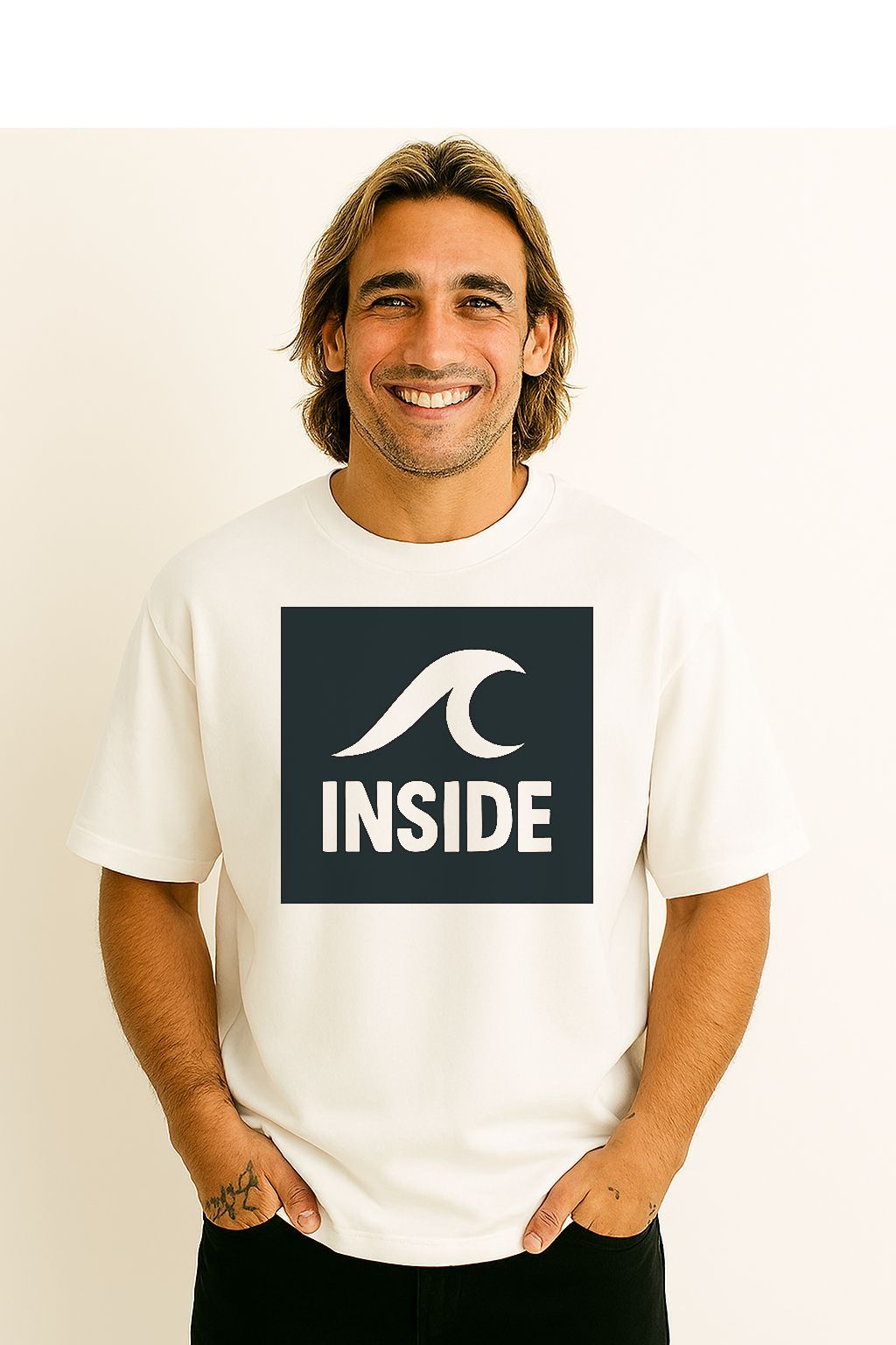 Homem vestindo uma camiseta branca com um logotipo quadrado azul, sorrindo. Mãos nos bolsos.