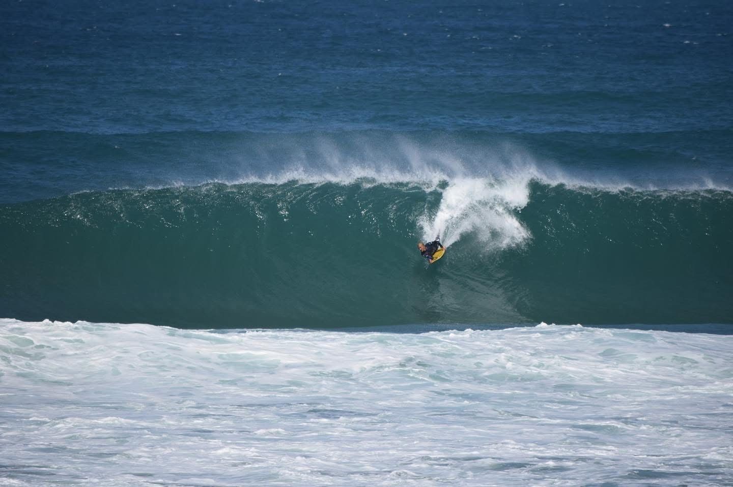 bodyboarder surfando dentro de uma grande onda turquesa, com espuma branca e oceano azul escuro ao fundo.