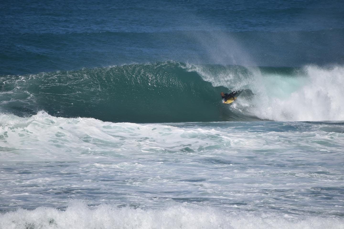 Bodyboarder em uma onda quebrando, cenário oceânico. Água e céu azuis, espuma branca.
