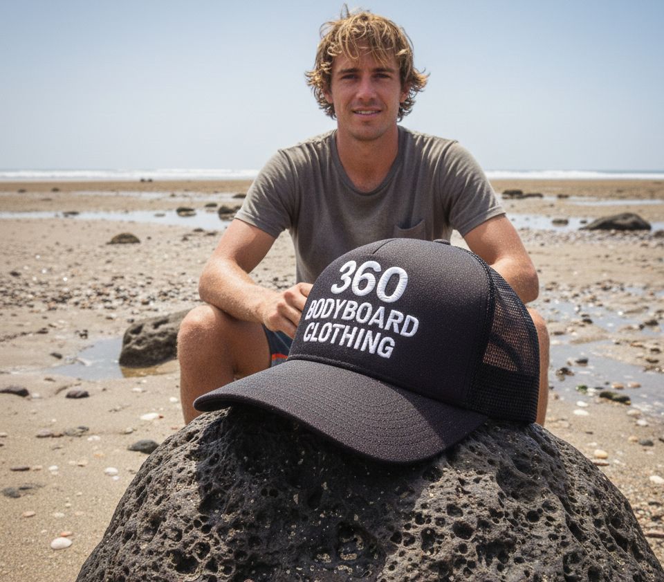 Homem sentado em rochas com um boné de bodyboard 360 na praia.