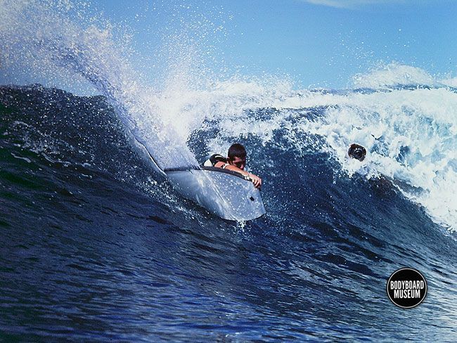 Pessoa praticando bodyboard dentro de uma onda quebrando; água azul, dia ensolarado.