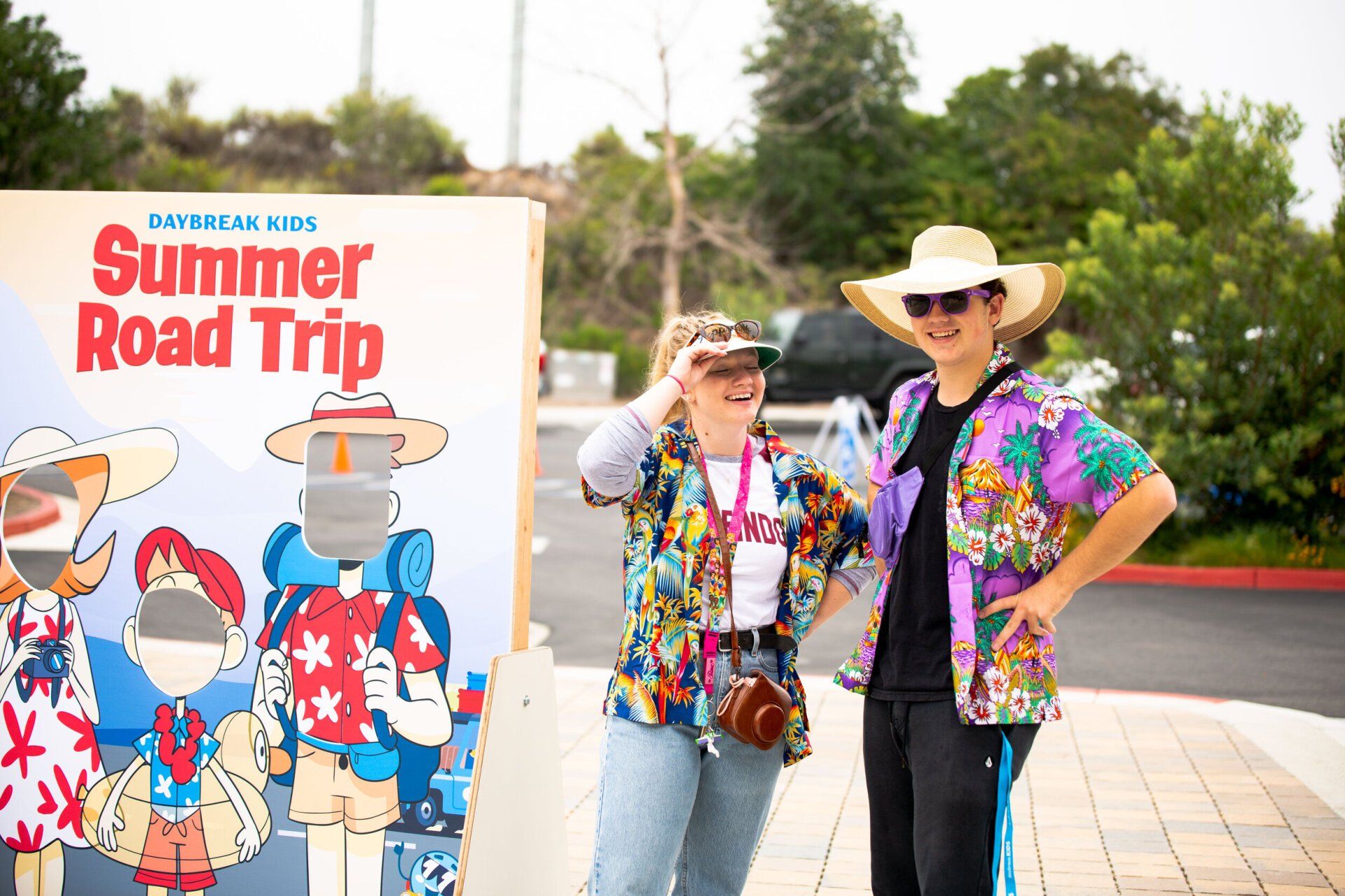 Two people are standing in front of a summer road trip sign.