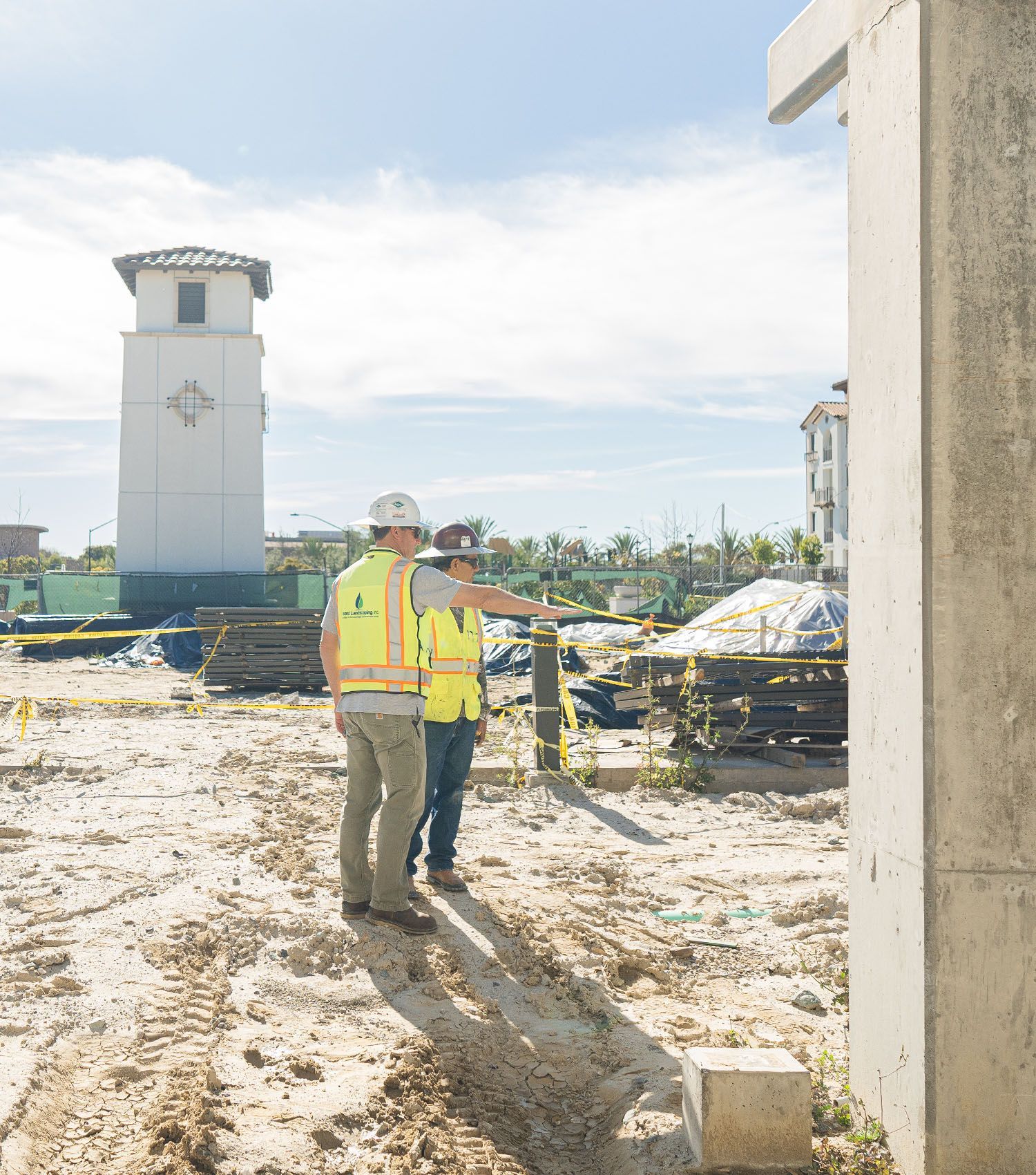 Two construction workers are standing in a dirt field with a clock tower in the background.