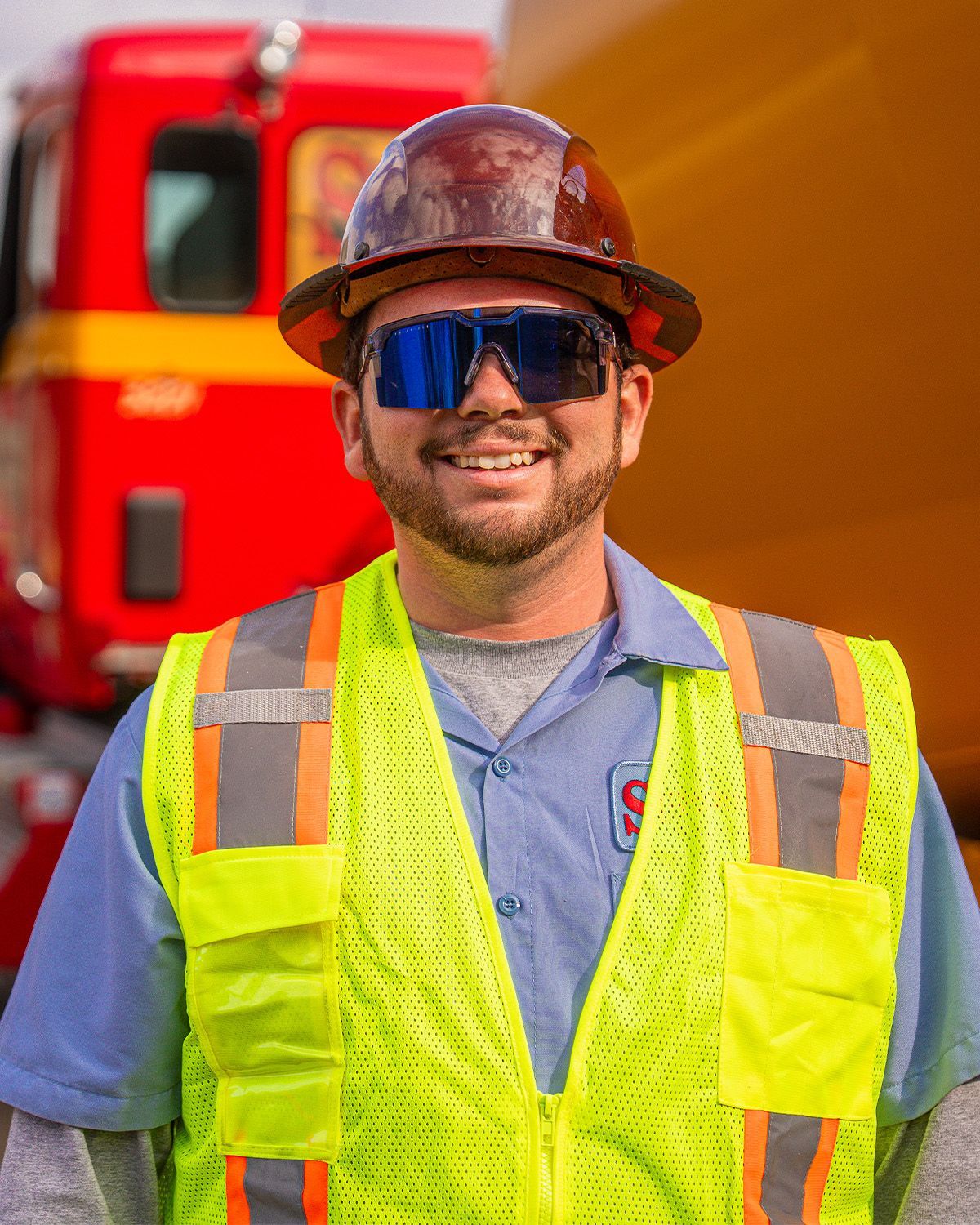 A man wearing a hard hat and sunglasses is smiling in front of a red truck.