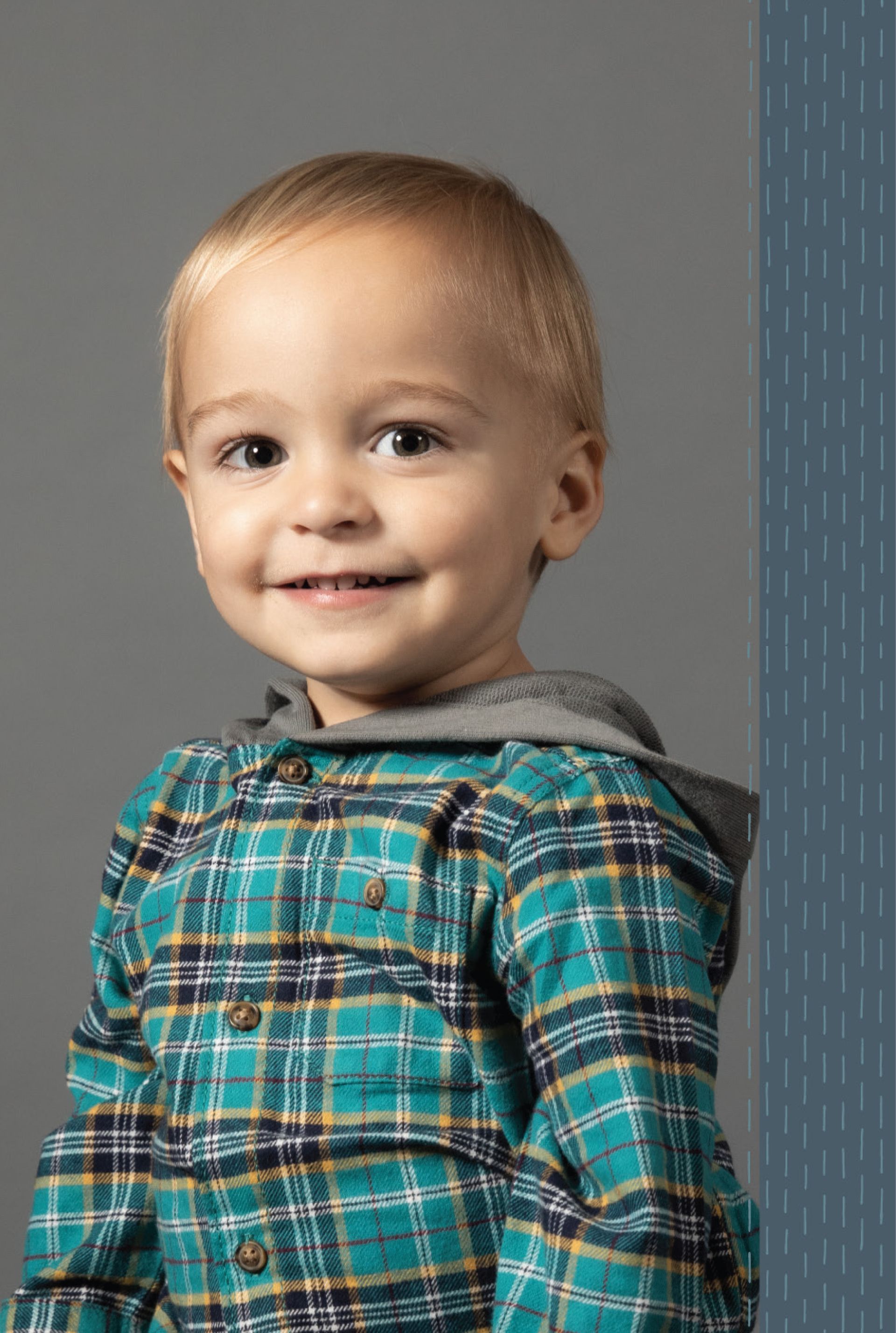 Blond toddler smiling, wearing a green plaid shirt with a gray hood, standing in front of a gray background.