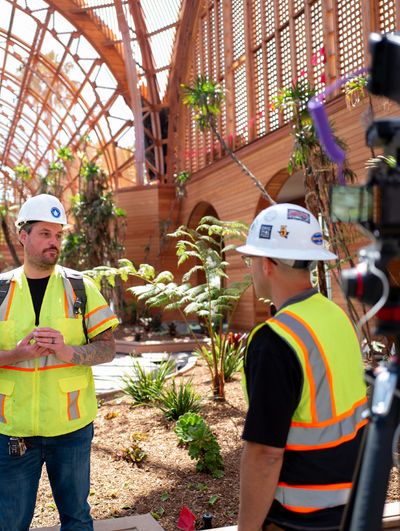 Two construction workers in hard hats and safety vests stand in a wooden greenhouse, one speaking to the other.