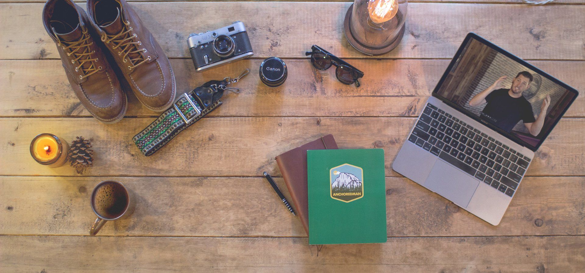 A wooden table with a laptop , a camera , sunglasses , a notebook , and a pair of boots.