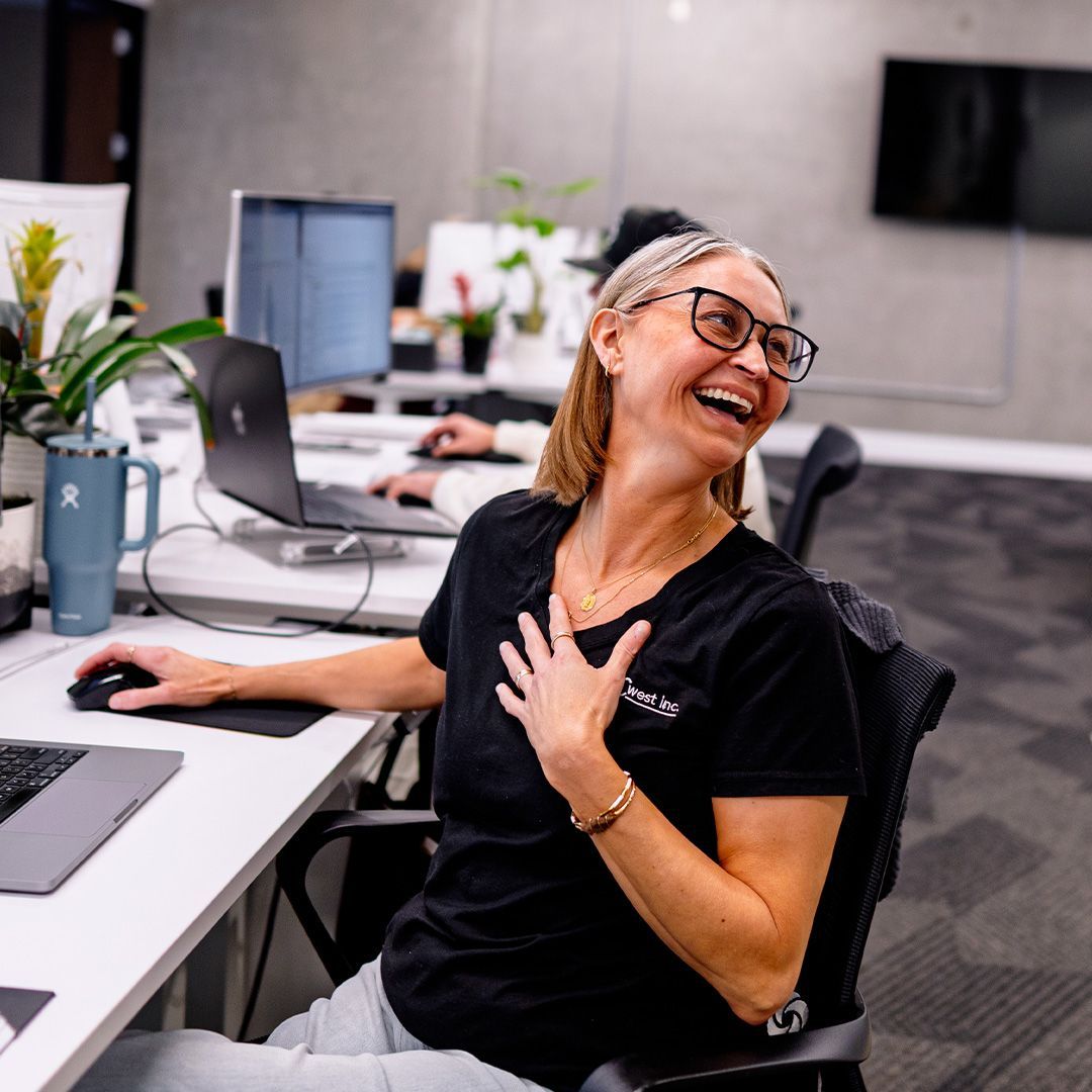 A smiling person wearing glasses and a black shirt sits at a desk in an office, laughing while placing a hand on their chest.