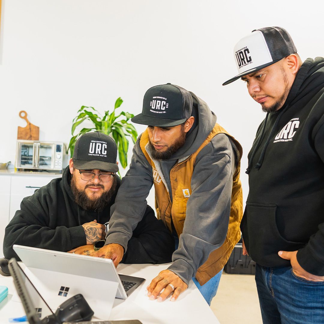 Three people wearing matching black-and-white hats look at a laptop screen in a brightly lit office setting.