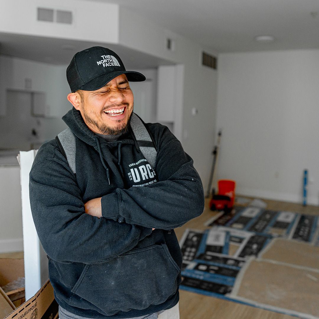 A person wearing a black cap and hoodie with arms crossed, laughing, standing in a room with light-colored flooring.