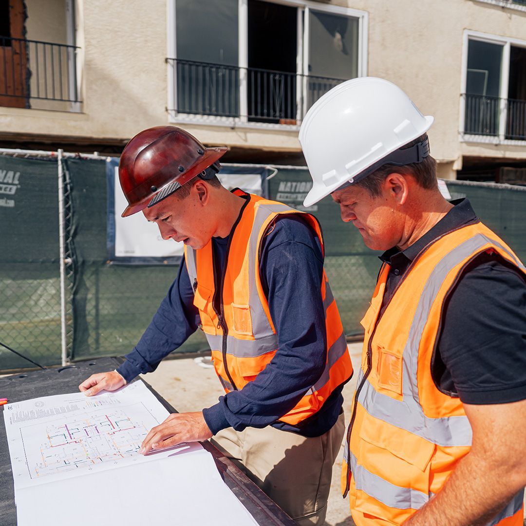 Two construction workers in high-visibility vests and hard hats review blueprints at an active construction site.