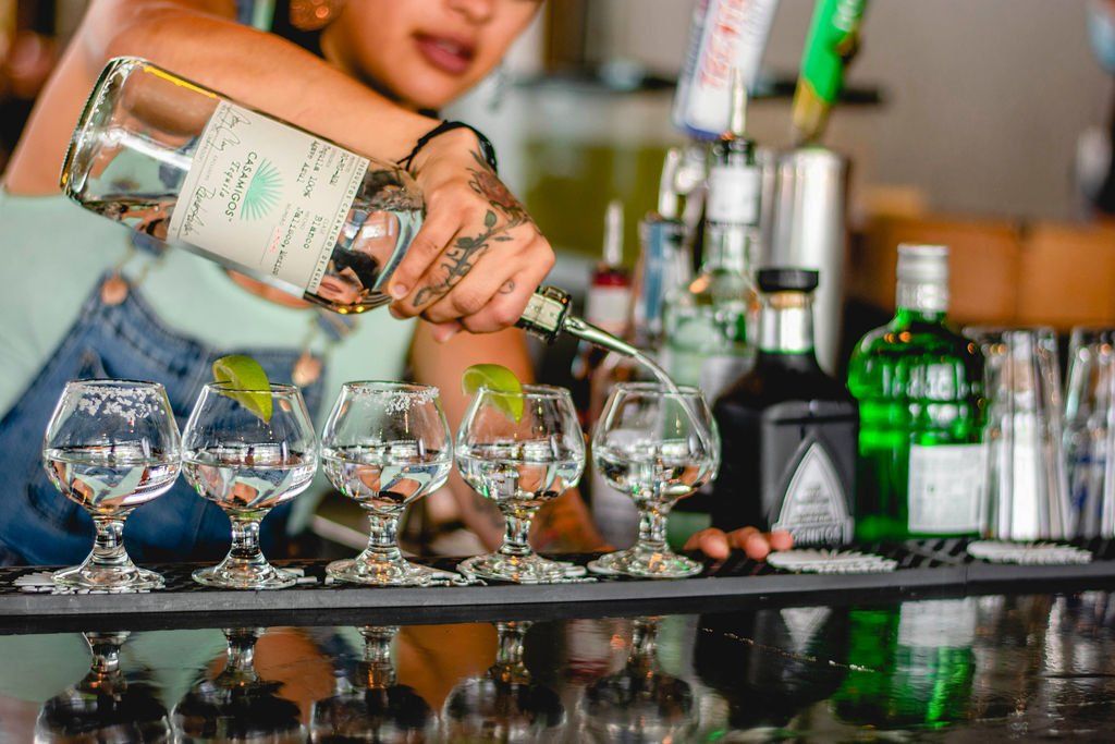 A bartender is pouring a drink into a row of wine glasses.