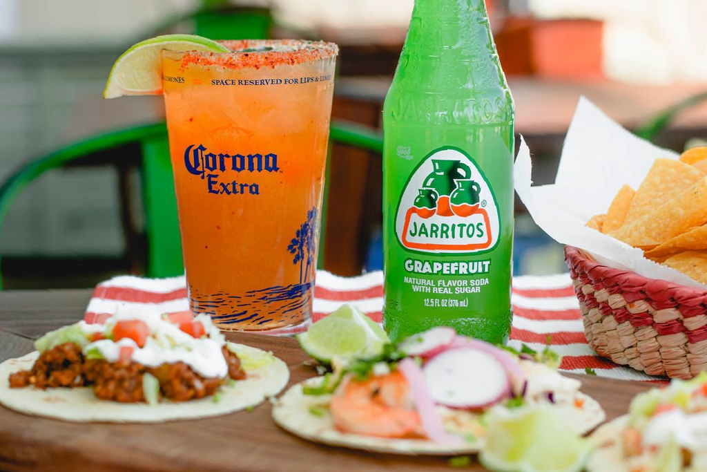 A table topped with tacos , a Jarritos drink , and a basket of chips.
