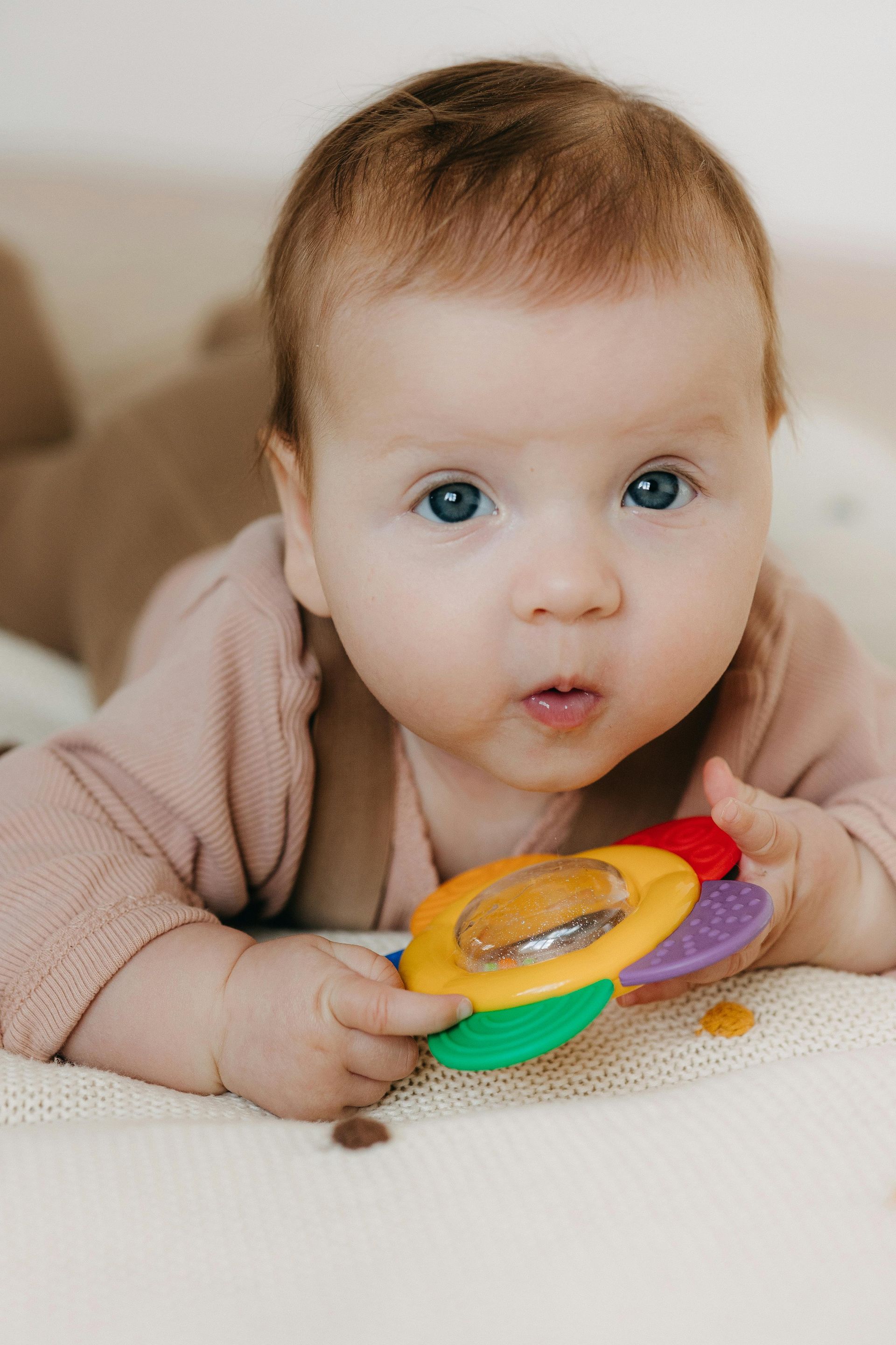 A baby lying on their stomach on a light surface, holding a colorful flower-shaped rattle.
