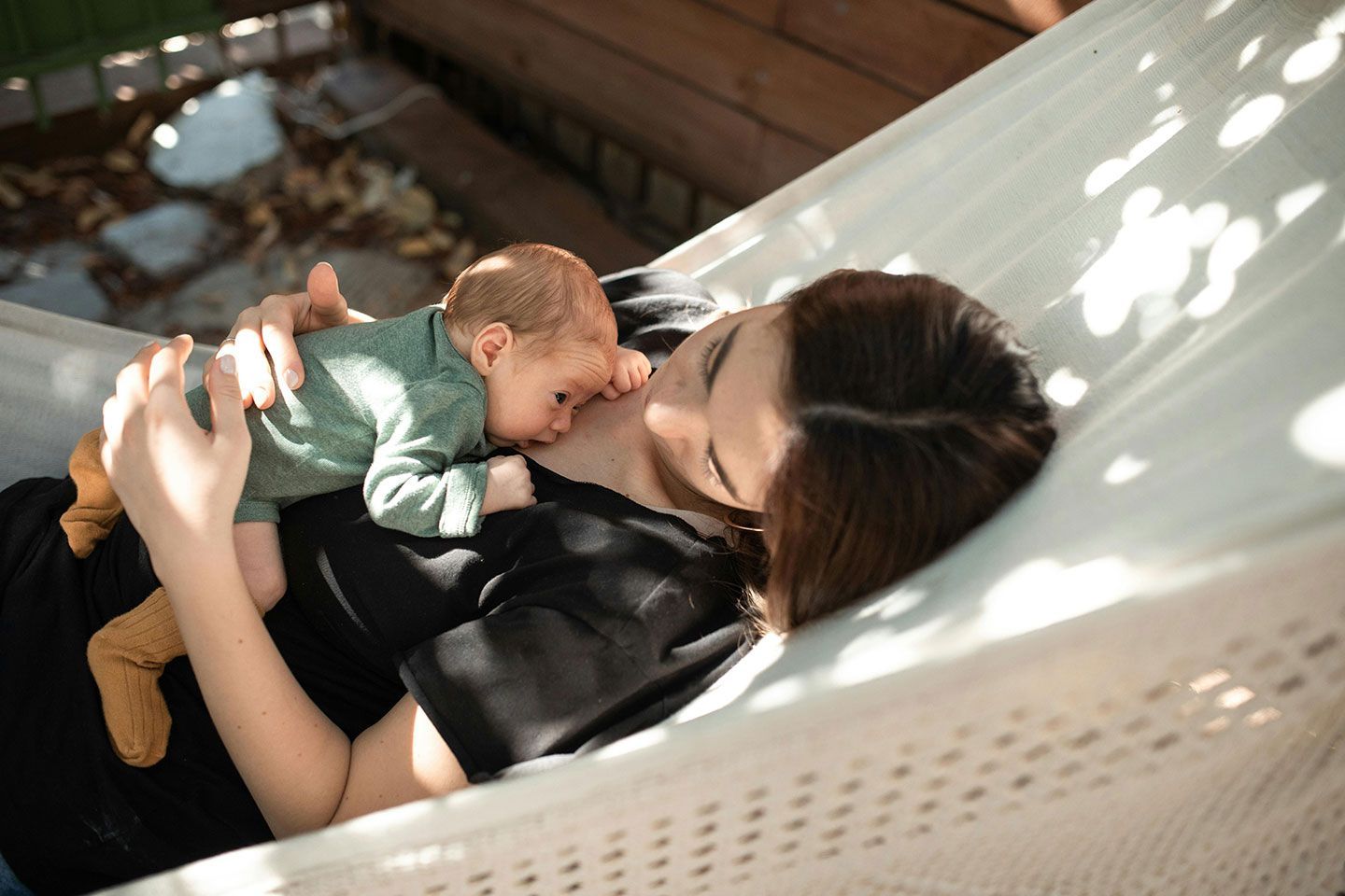 Woman and newborn baby cuddle in a hammock outdoors.