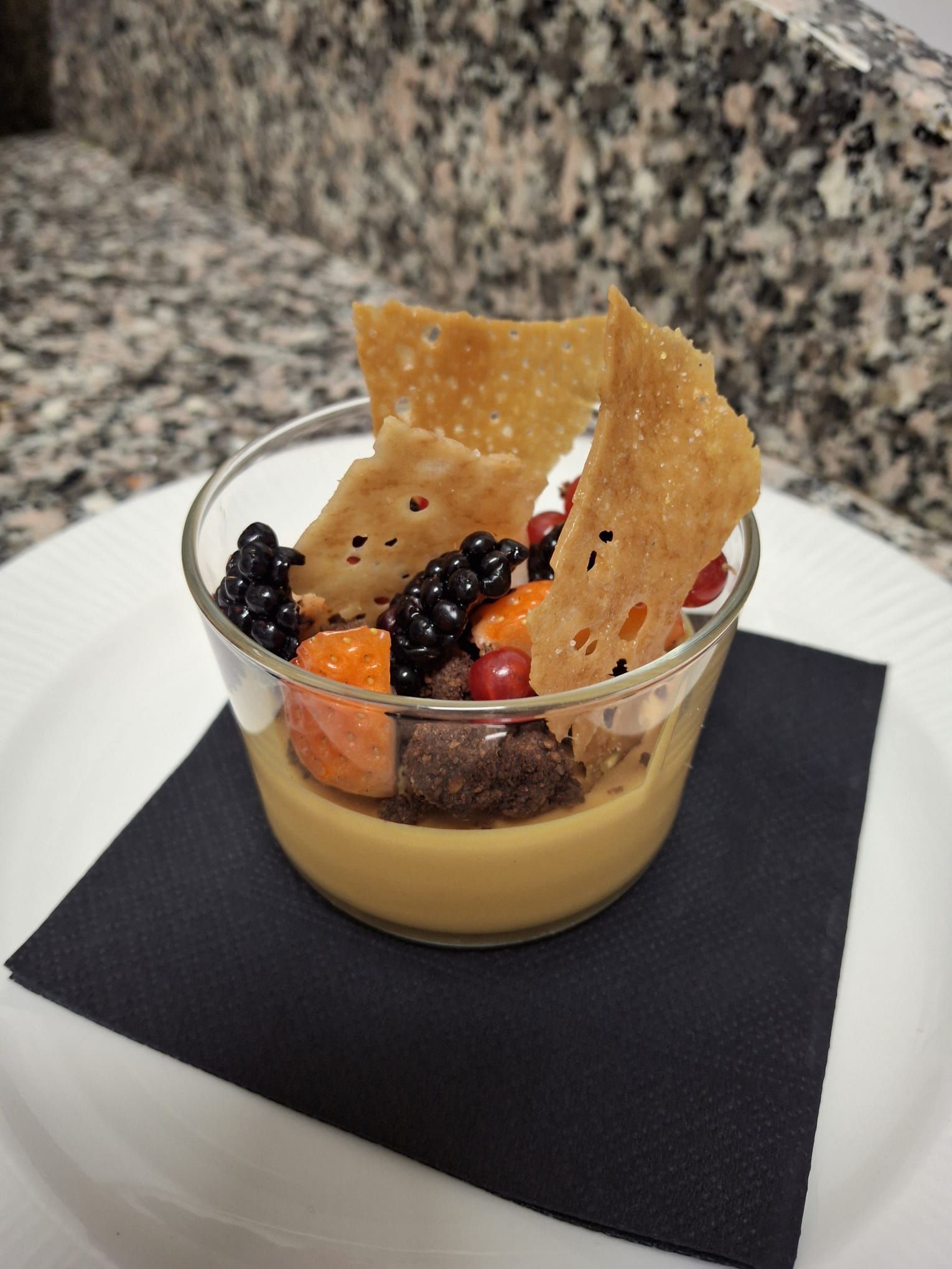 Dessert in bicchiere: mousse al caramello a strati, frutti di bosco, cioccolato e pasta croccante su un quadrato nero, adagiato su un piatto bianco.