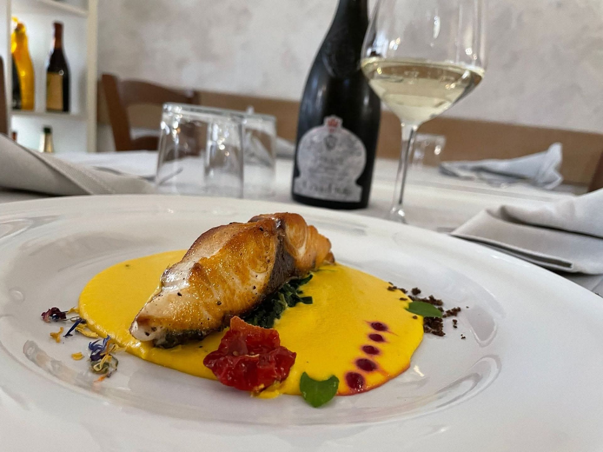 Pesce scottato in padella con salsa gialla, servito con un bicchiere di vino e una bottiglia su una tovaglia bianca.