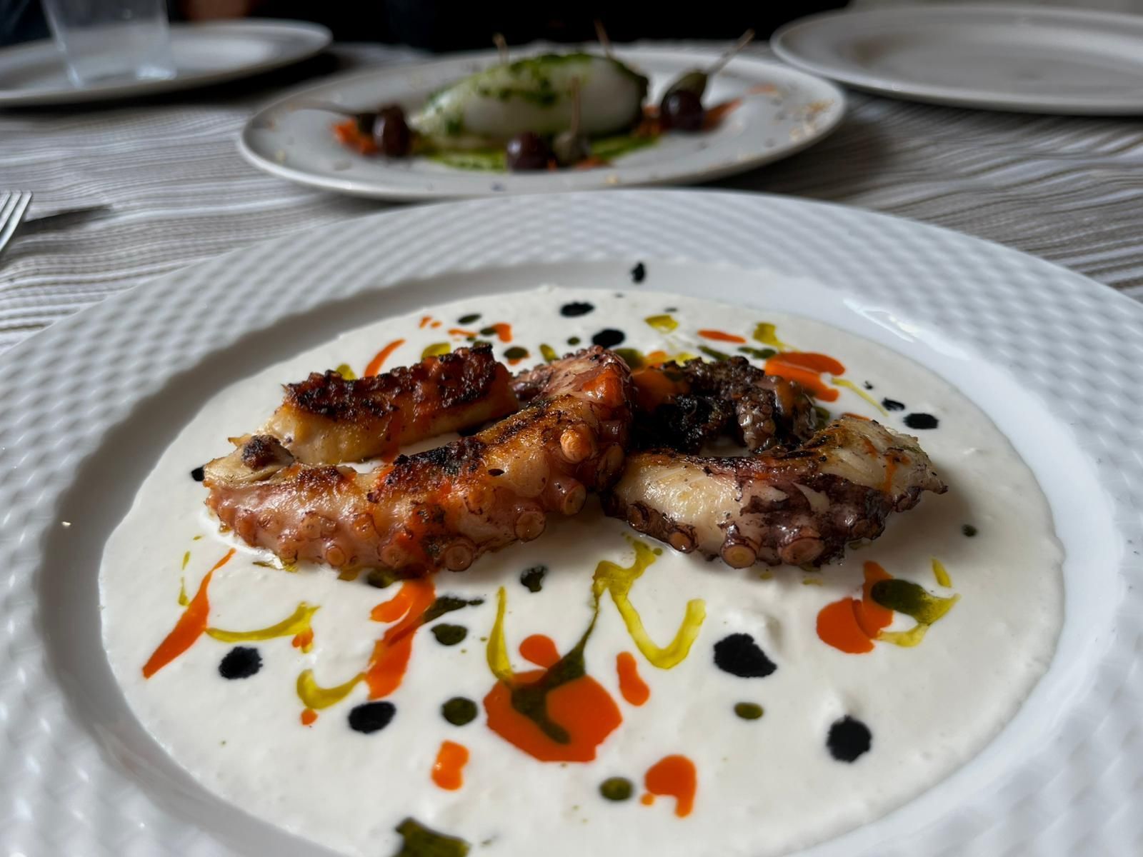 Polpo alla griglia su salsa bianca con gocce colorate, servito su un piatto bianco.