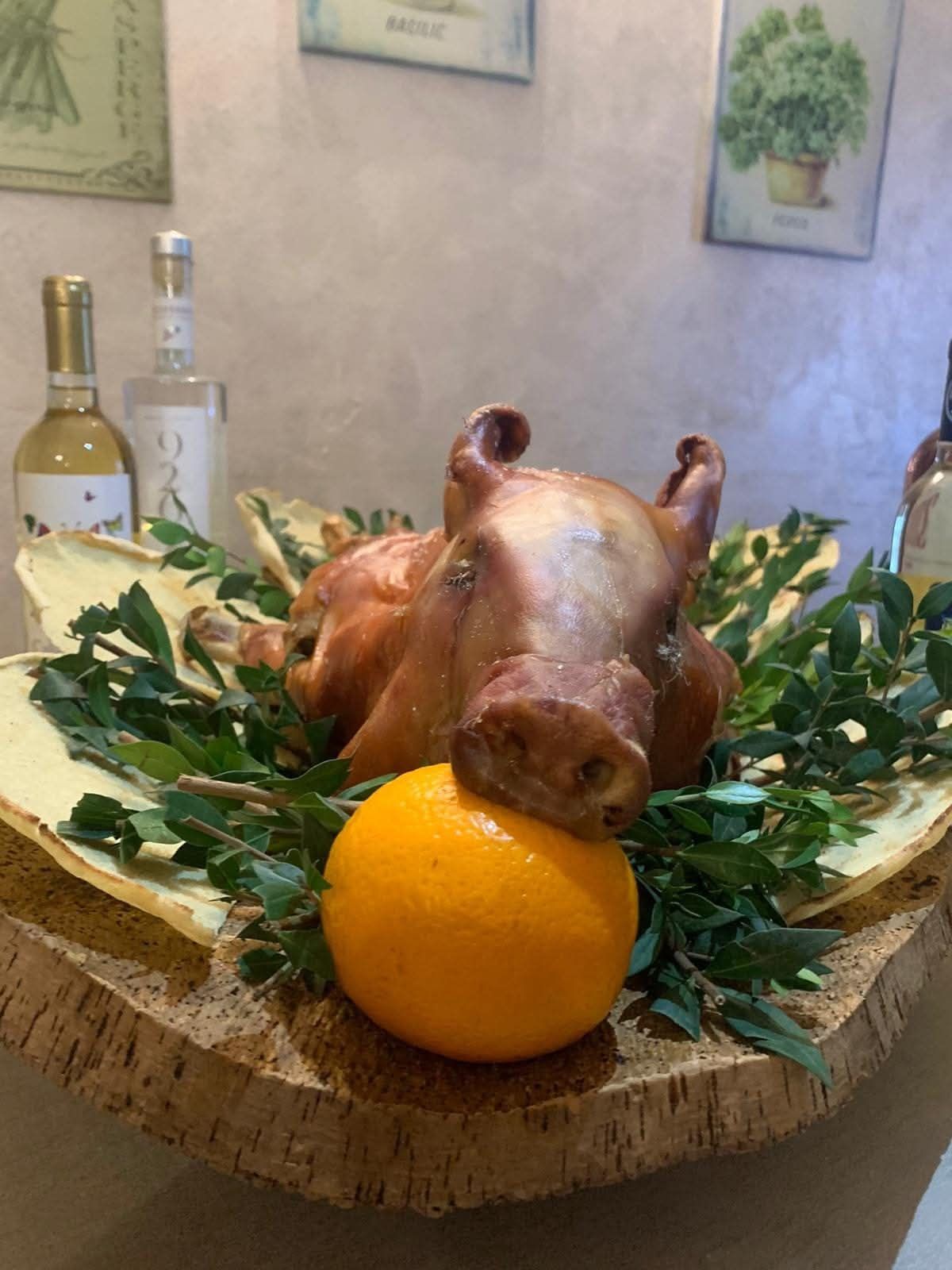 Testa di maiale arrosto su un vassoio di legno con verdure e un'arancia. Bottiglie di vino sullo sfondo.