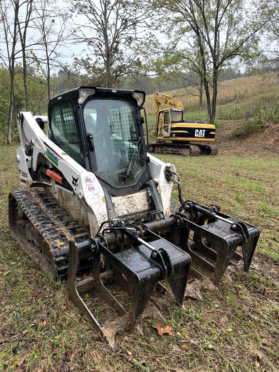A white Bobcat compact track loader with a grapple attachment parked in a field, with a yellow CAT excavator behind it.
