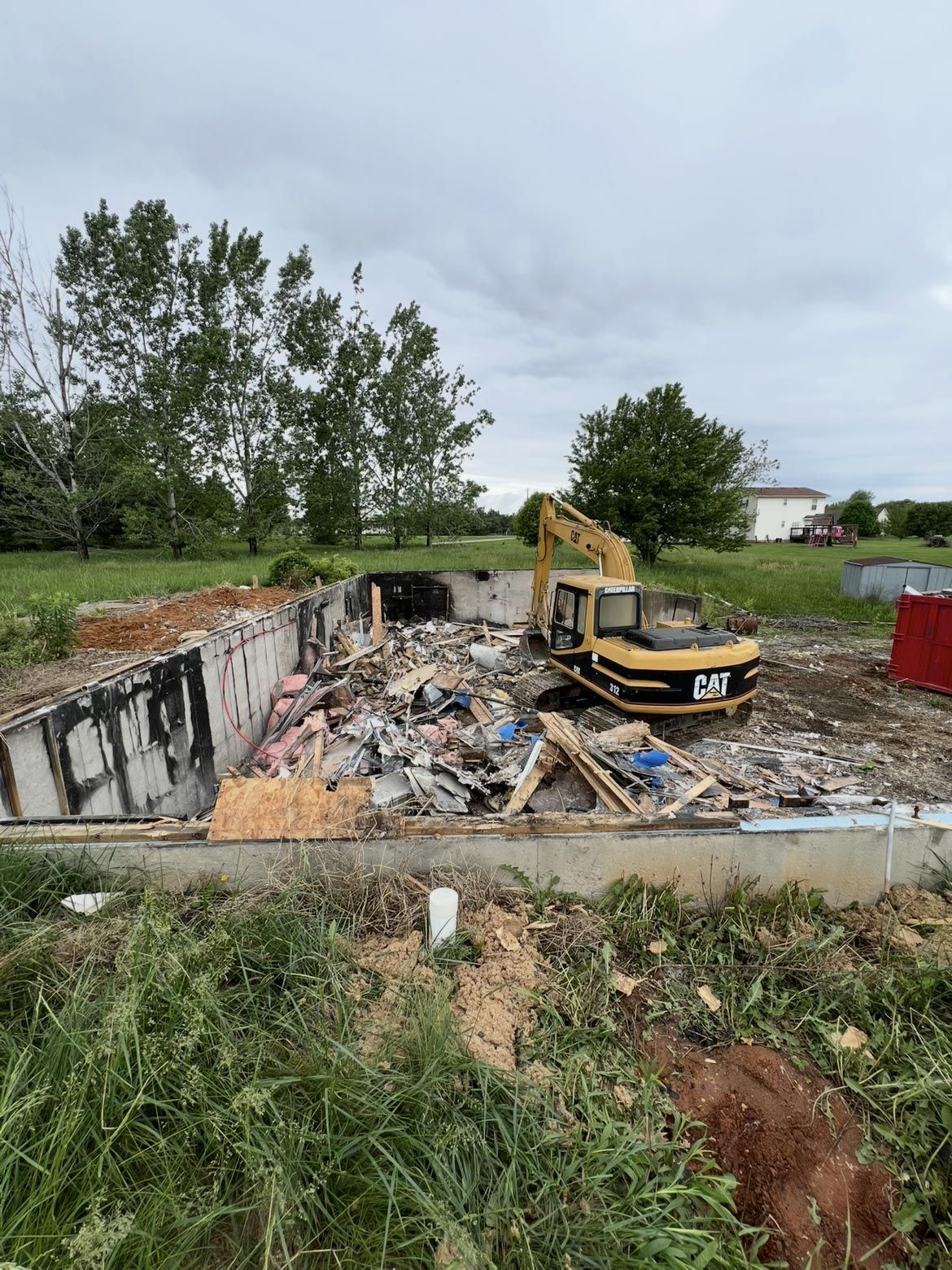 A yellow excavator works inside a concrete foundation filled with demolition debris in a grassy, open outdoor setting.