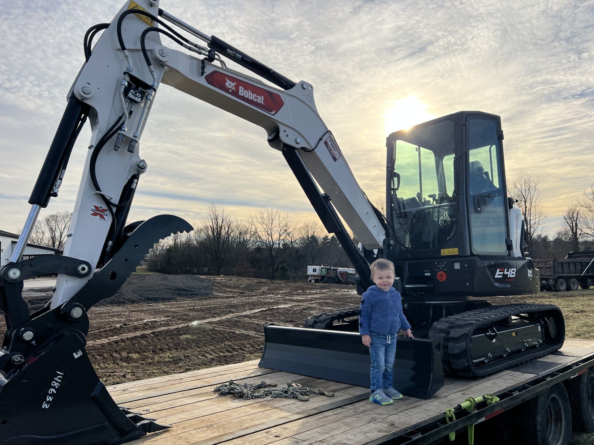 A small child stands on a flatbed trailer next to a white and black Bobcat excavator at sunset.