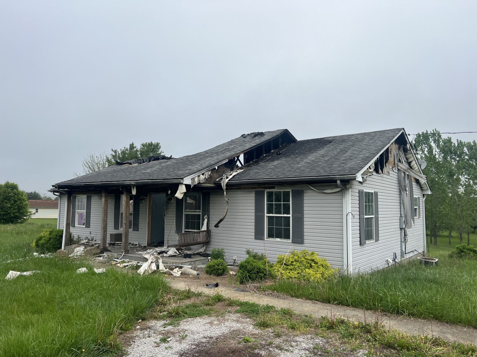 A single-story, light-colored house with significant fire damage to the roof and front porch, set on a grassy lot.