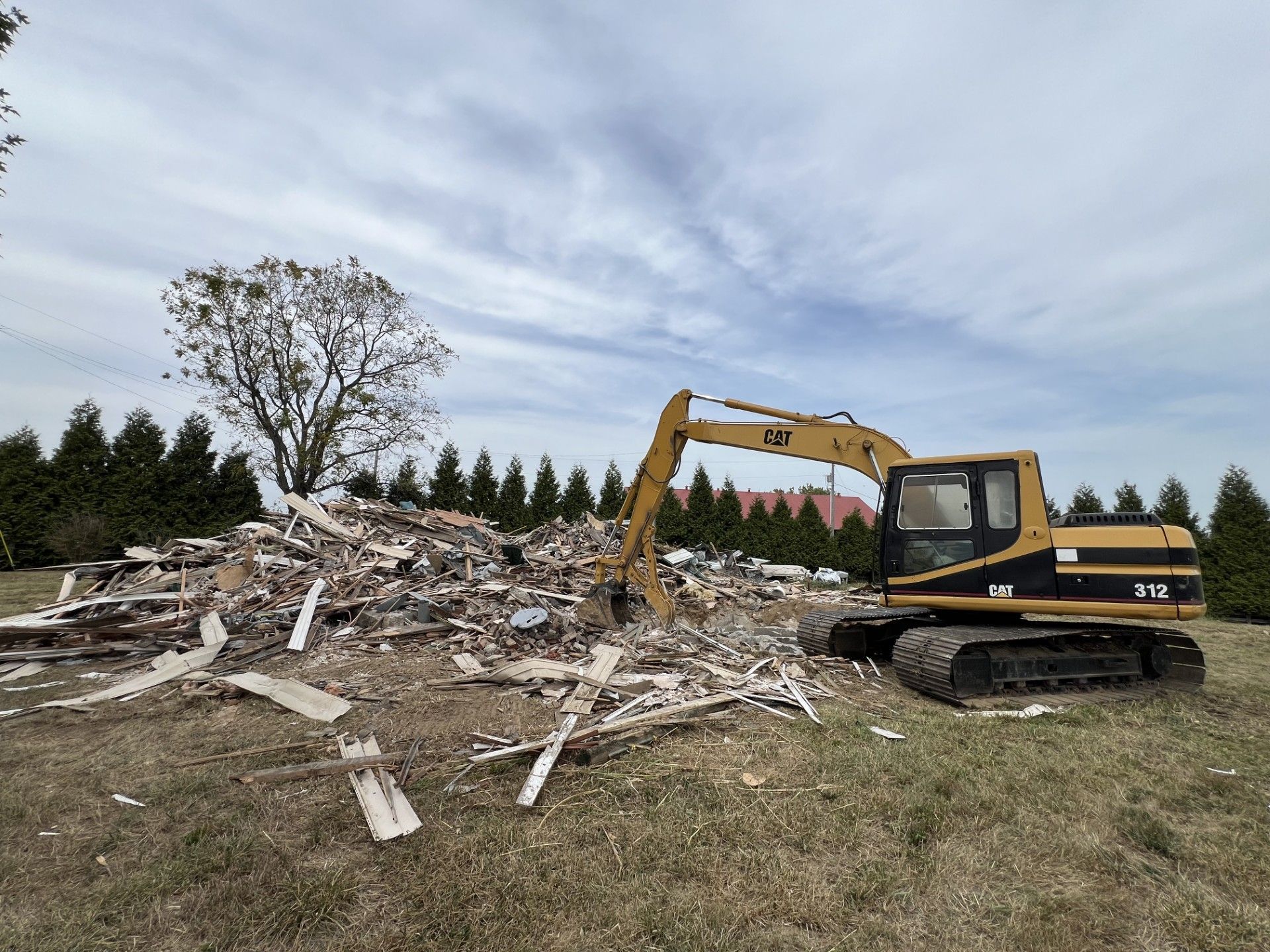 A yellow CAT excavator sits on a grassy field next to a large pile of debris from a demolished building.