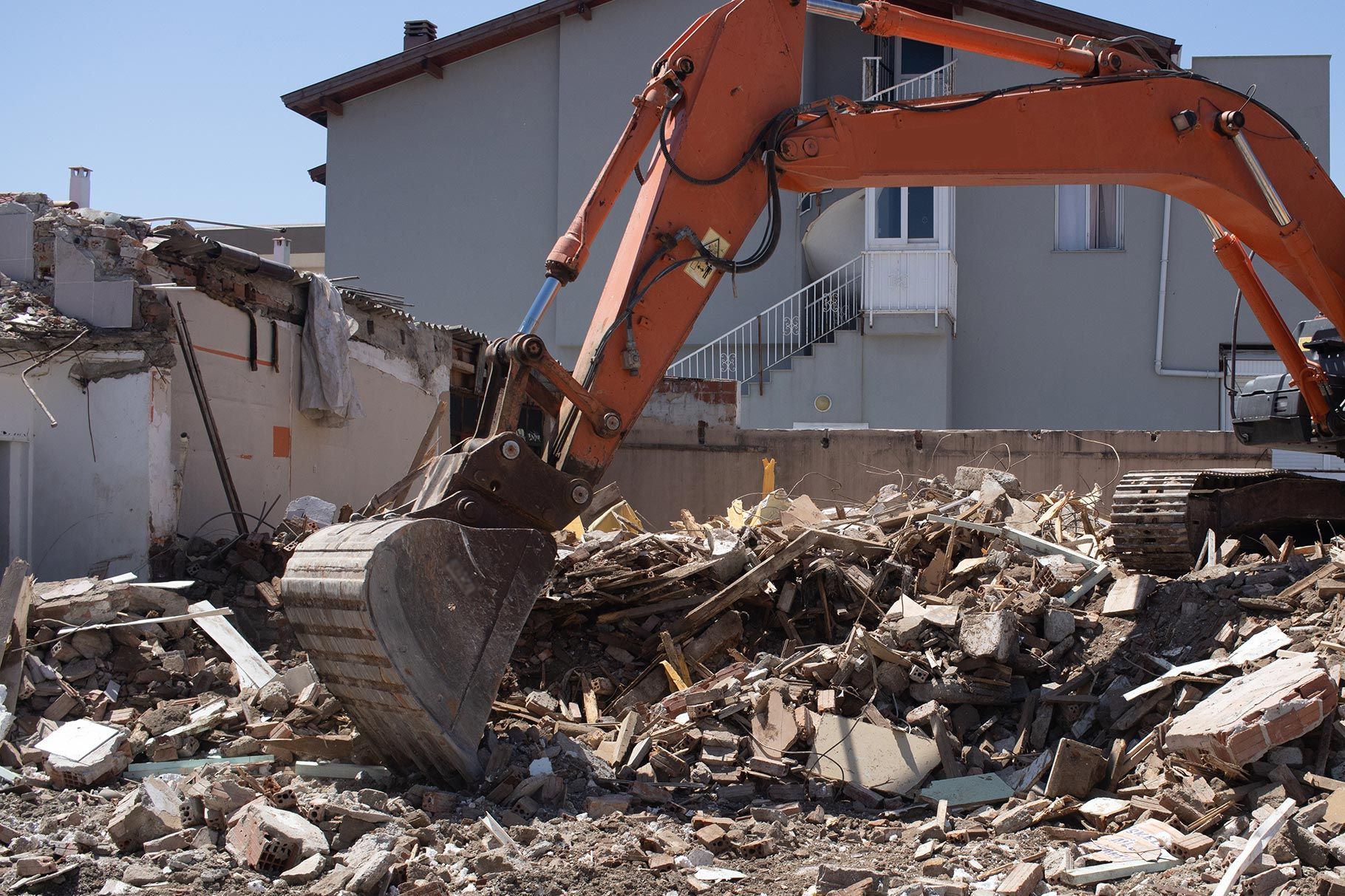 An orange excavator sits atop a pile of concrete rubble at a demolition site in front of a building. An orange excavator sits atop a pile of concrete rubble at a demolition site in front of a building.