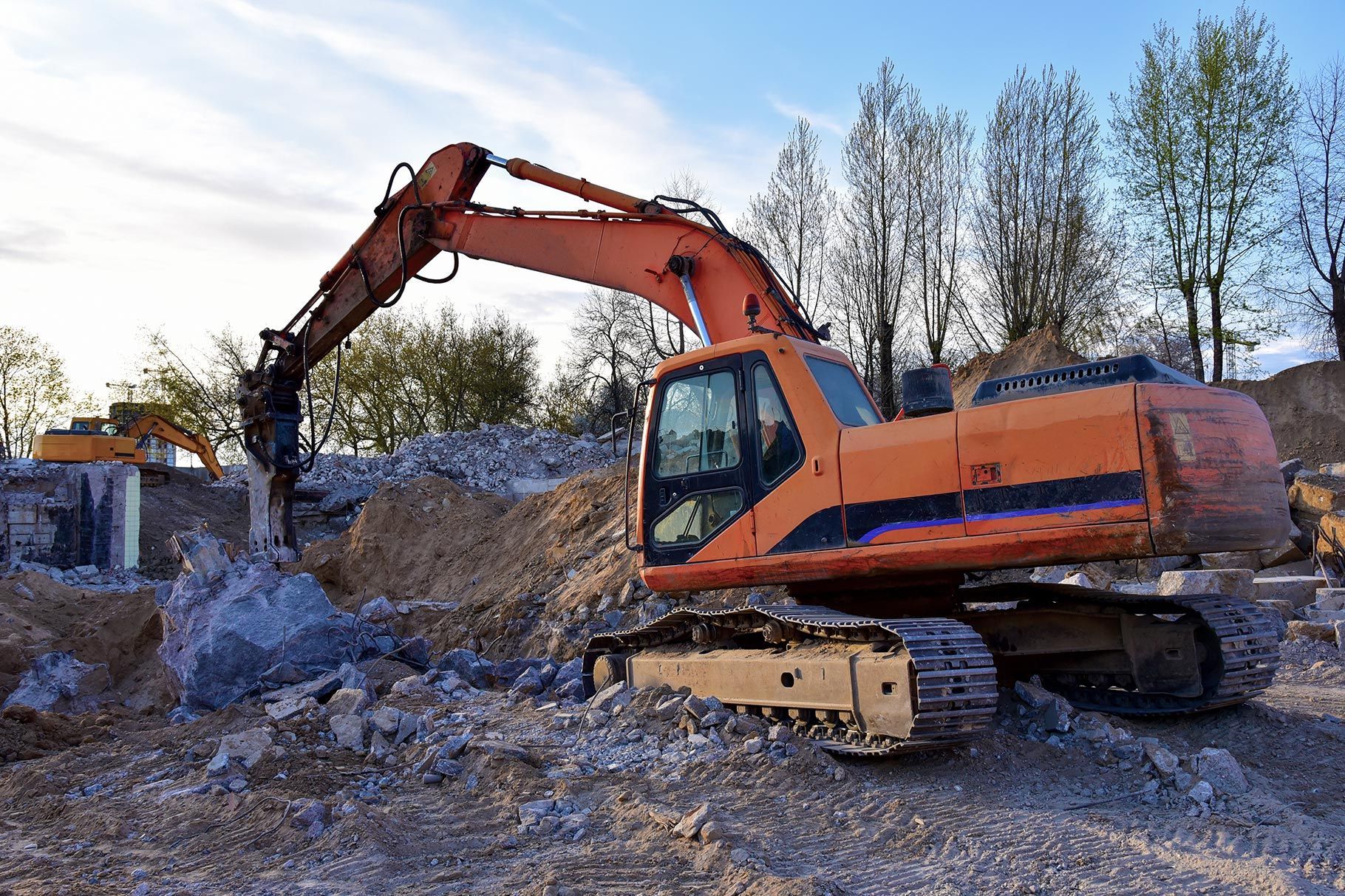 An orange excavator with a hydraulic breaker attachment works on a demolition site, breaking up piles of concrete rubble. An orange excavator with a hydraulic breaker attachment works on a demolition site, breaking up piles of concrete rubble.