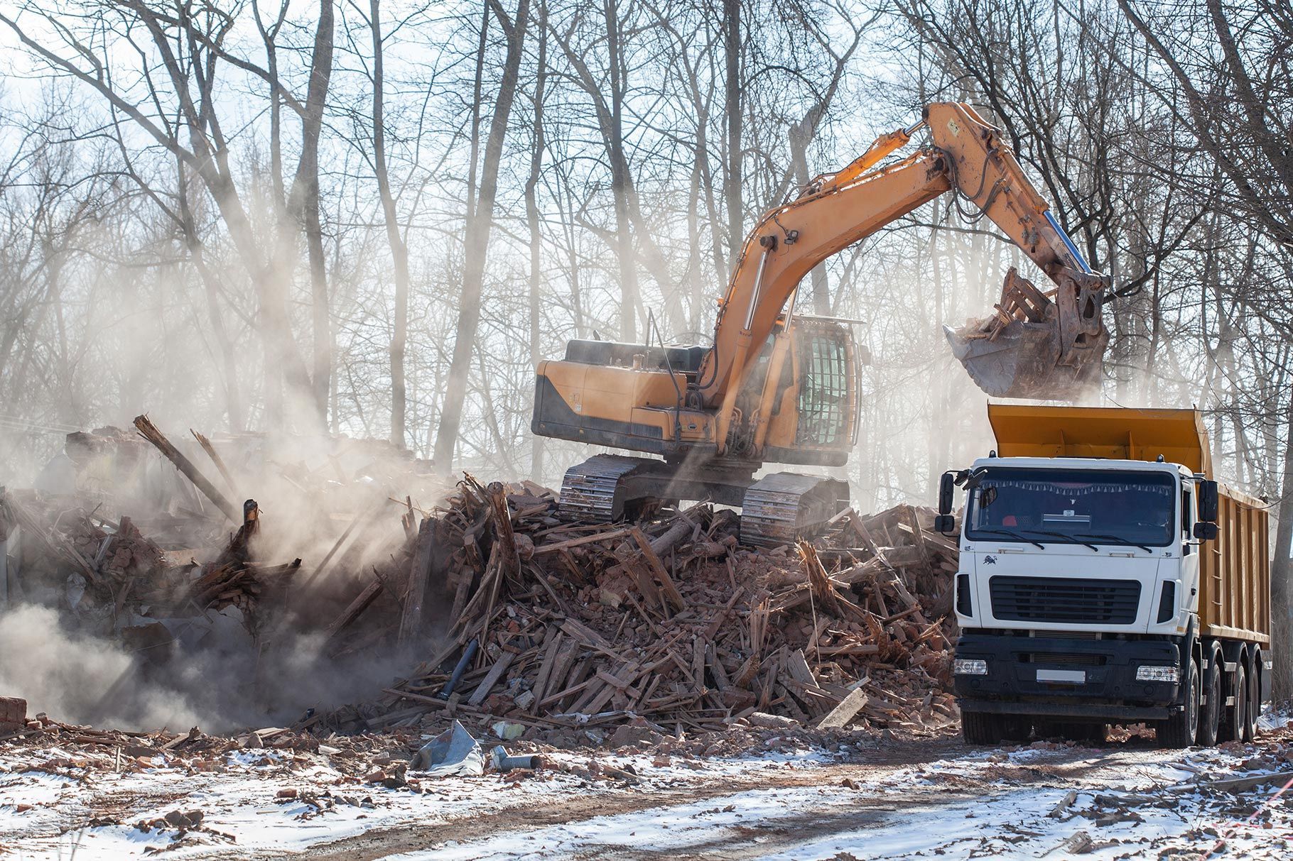 A yellow excavator loads debris into a white dump truck amidst a dusty, snow-covered landscape with bare trees. A yellow excavator loads debris into a white dump truck amidst a dusty, snow-covered landscape with bare trees.