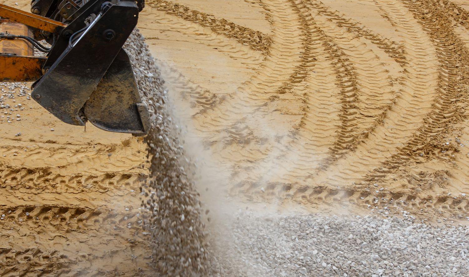 An excavator bucket pours gravel onto a sandy construction site with visible tire tracks. An excavator bucket pours gravel onto a sandy construction site with visible tire tracks.