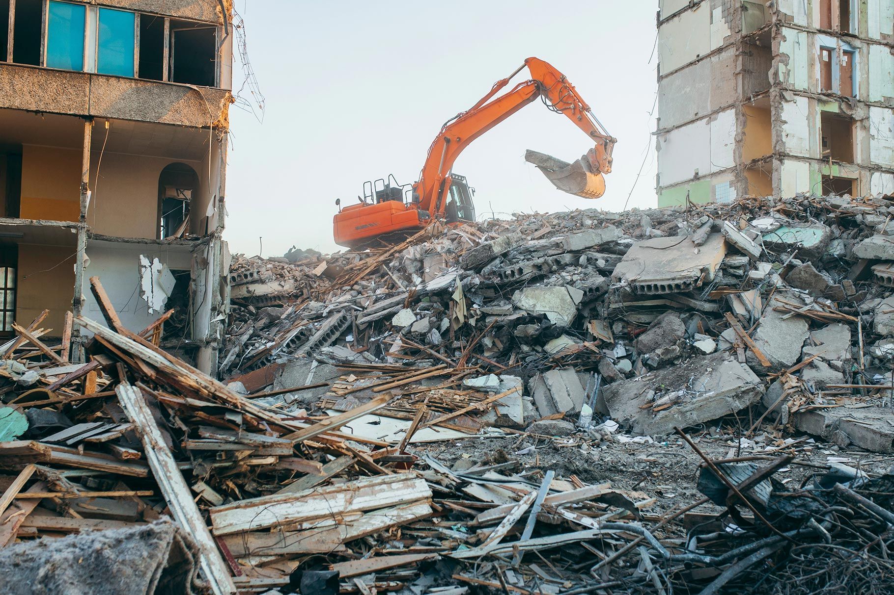 An orange excavator works on a large pile of rubble between two partially demolished multistory buildings. An orange excavator works on a large pile of rubble between two partially demolished multistory buildings.
