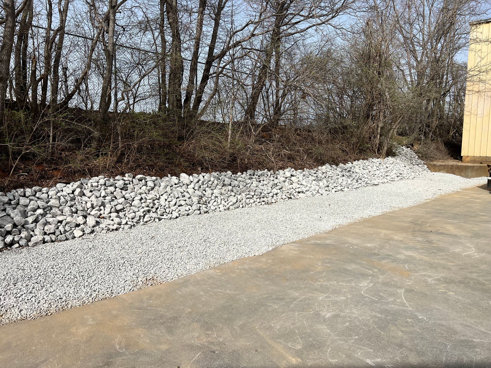A gravel driveway alongside a low retaining wall made of large, light-colored stones, set against a backdrop of bare trees.