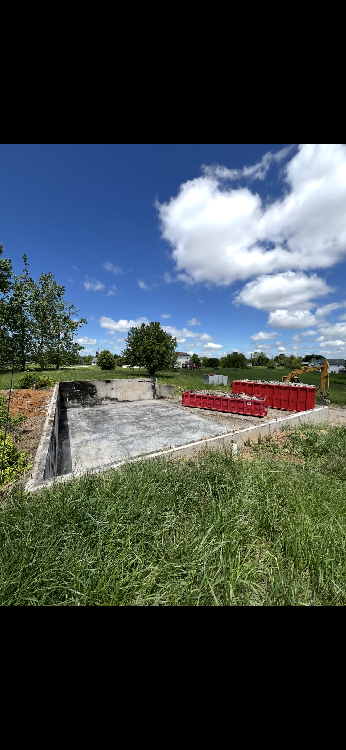 An outdoor construction site featuring a concrete foundation and red debris bins under a bright blue, cloudy sky.