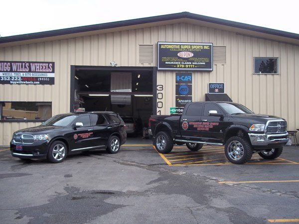 Two black SUVs parked in front of an automotive shop. The shop is tan with a large open bay door.