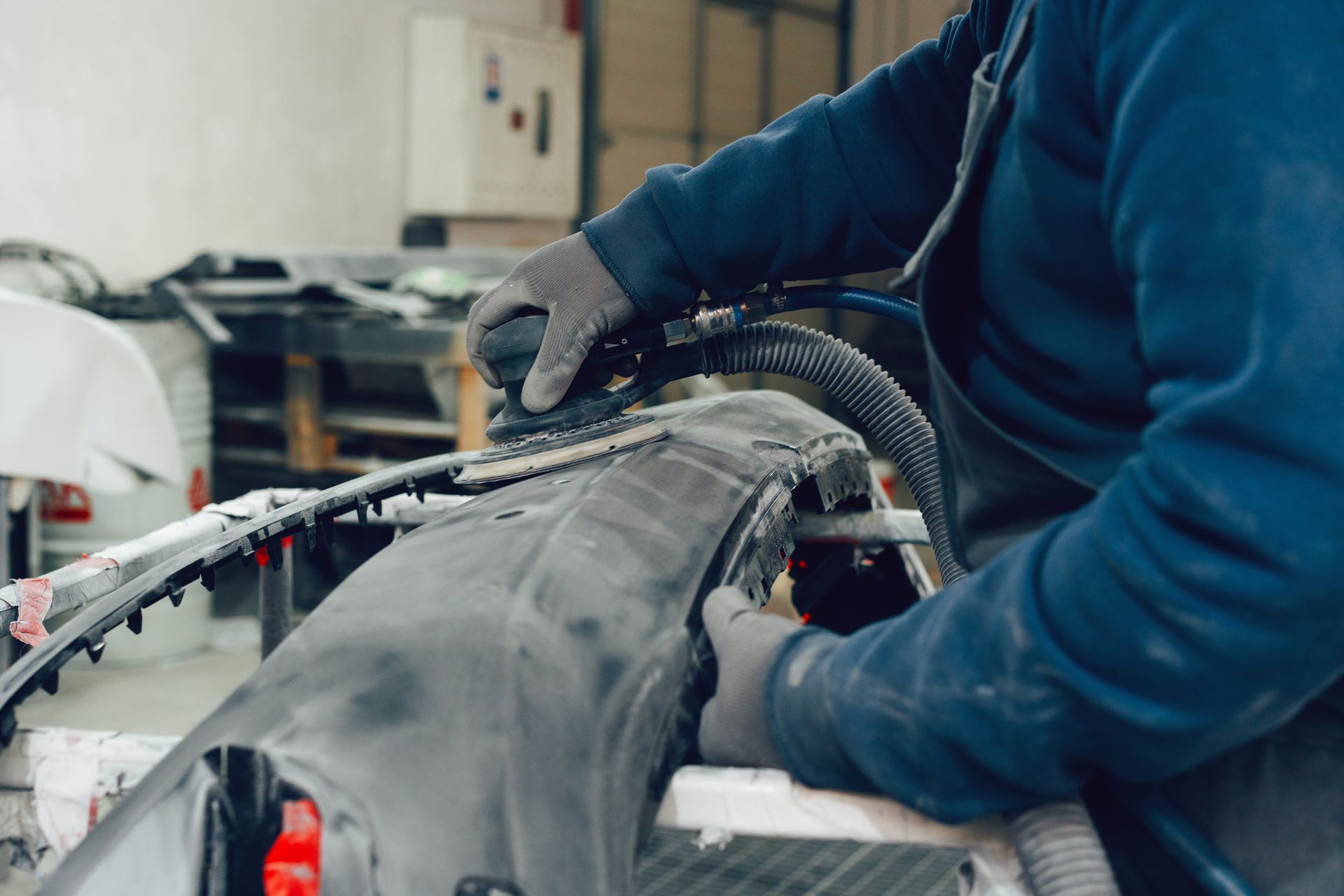 A mechanic wearing blue clothing and gloves is repairing a bumper at an auto collision repair shop.
