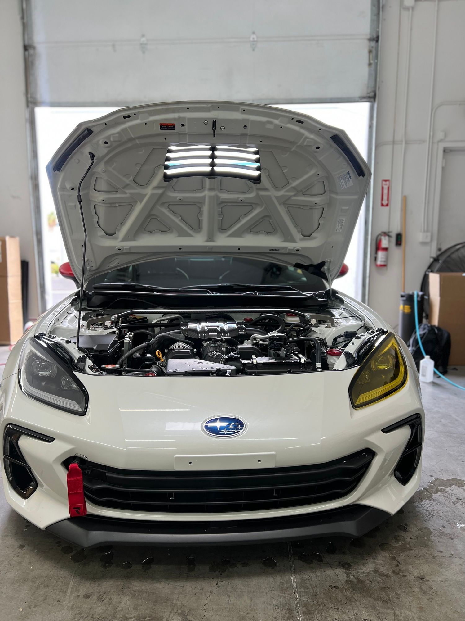 A white subaru brz with the hood open is parked in a garage.