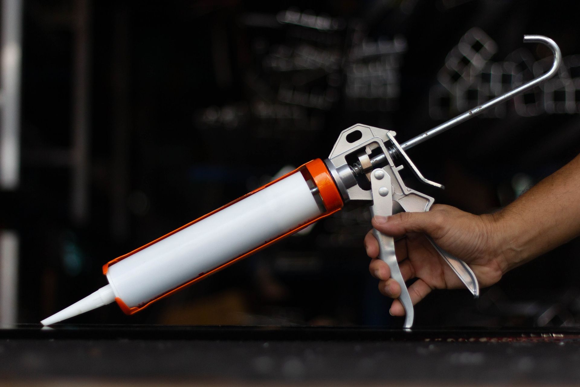 Hand holding a caulking gun with a white and orange tube, ready to dispense sealant.