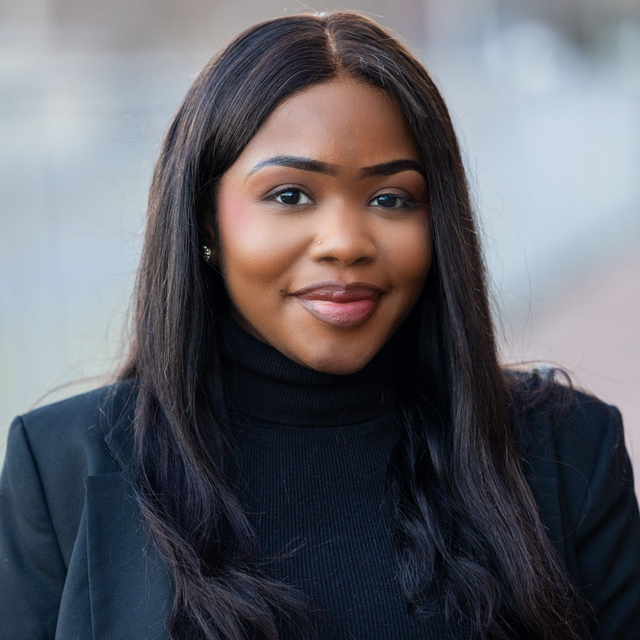 A headshot of April Vaughn wearing a black turtleneck and blazer, smiling softly against a blurred background.