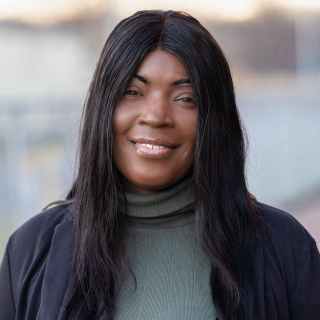 A headshot of Dee Williams wearing an olive-green turtleneck and black jacket, against a blurred background.